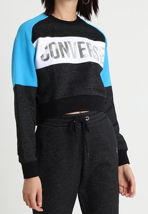Vrouw draagt een zwart-blauwe cropped trui met lange mouwen met tekst "CONVERSE" en bijpassende zwarte broek met trekkoord.