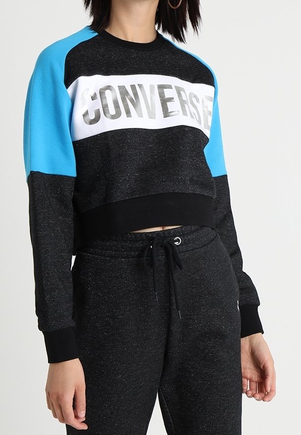 Femme portant un sweat-shirt court à manches longues noir et bleu avec le texte "CONVERSE" et un pantalon noir à cordon assorti.