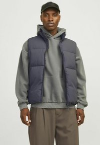 Námornícky puffer vest s predným zipsom, s vysokým golierom a dvoma bočnými vreckami, nosený cez šedú mikinu a béžové nohavice.