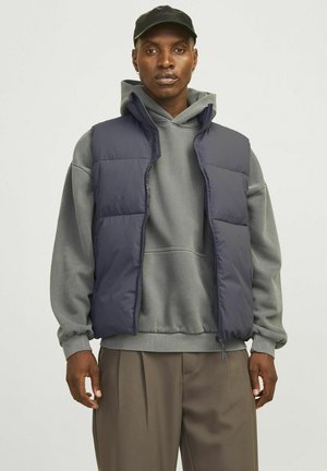 Námornícky puffer vest s predným zipsom, s vysokým golierom a dvoma bočnými vreckami, nosený cez šedú mikinu a béžové nohavice.