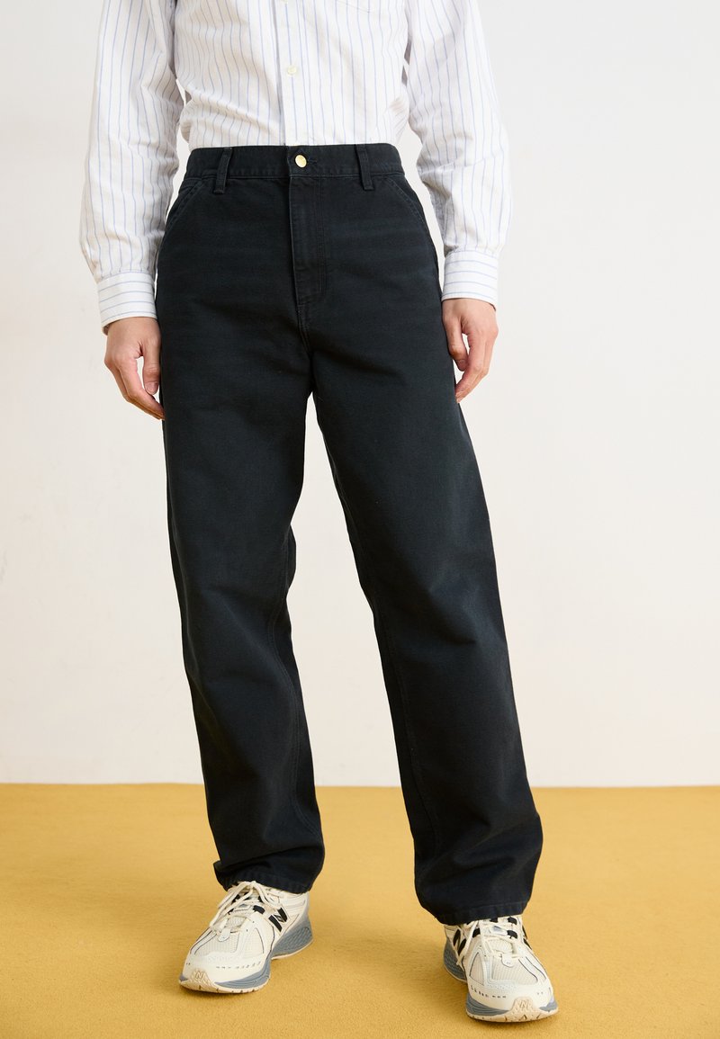 Black cotton trousers with a relaxed fit, featuring a front button closure and side pockets, paired with light-colored athletic shoes.