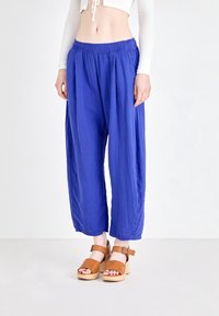 Pantalon large bleu léger plissé avec une taille élastique, assorti à des sandales à talons en bois beige. Le tissu a une finition texturée.