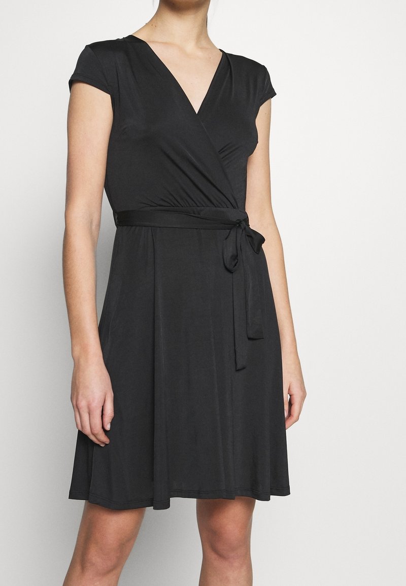 Femme portant une robe portefeuille noire à manches courtes nouée avec une ceinture, debout devant un fond clair uni.