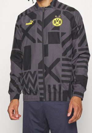 Czarna i szara kurtka zapinana na zamek błyskawiczny z geometrycznym wzorem, z żółtymi logo Puma i BVB 09, noszona przez osobę w granatowych spodniach.