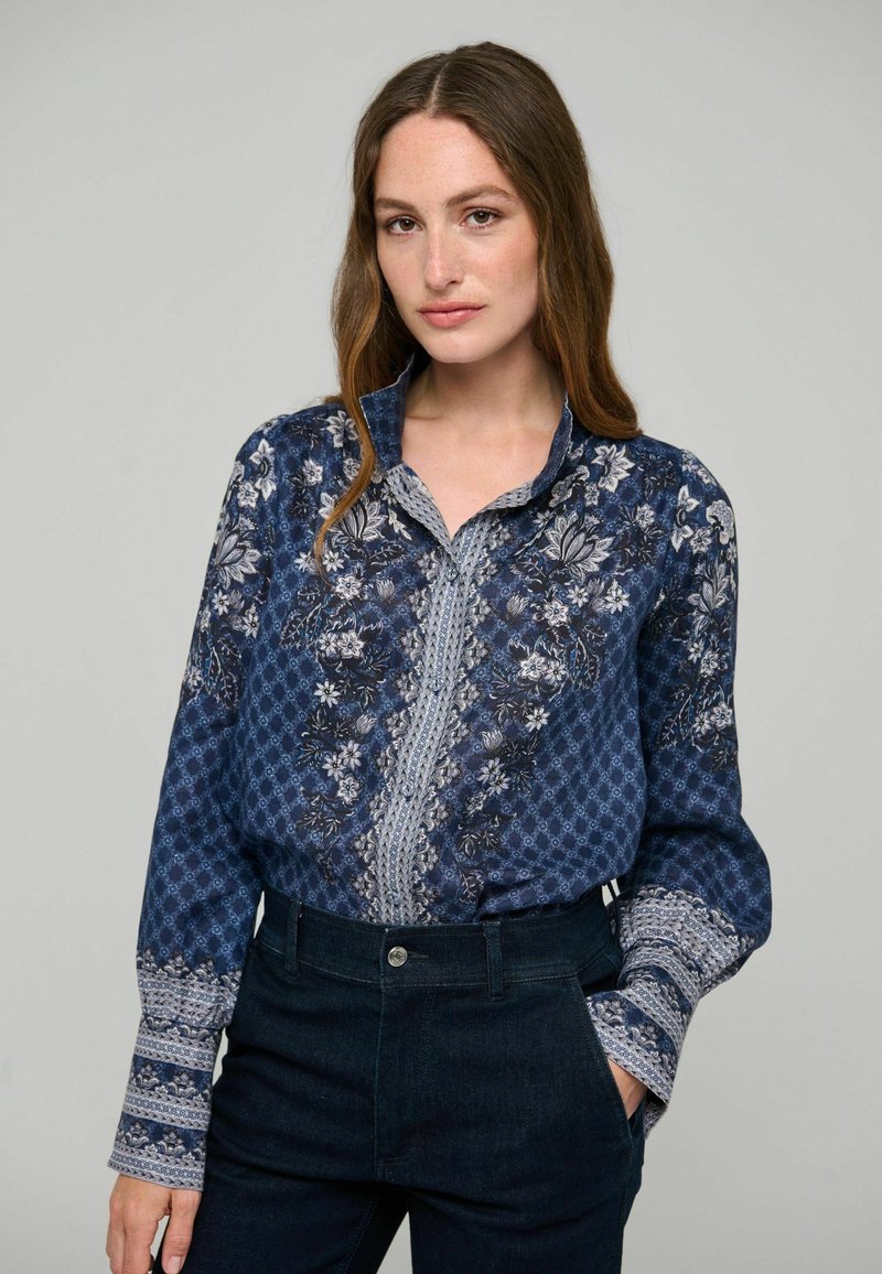Femme aux longs cheveux bruns portant un chemisier bleu à motif floral rentré dans un pantalon sombre taille haute, debout avec une main dans la poche.