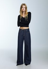 Široke hlače od tamnog trapera s kontrastnim detaljima šavova i crnim kožnim remenom. Model nosi crnu majicu s dugim rukavima, stoji s prekriženim rukama.