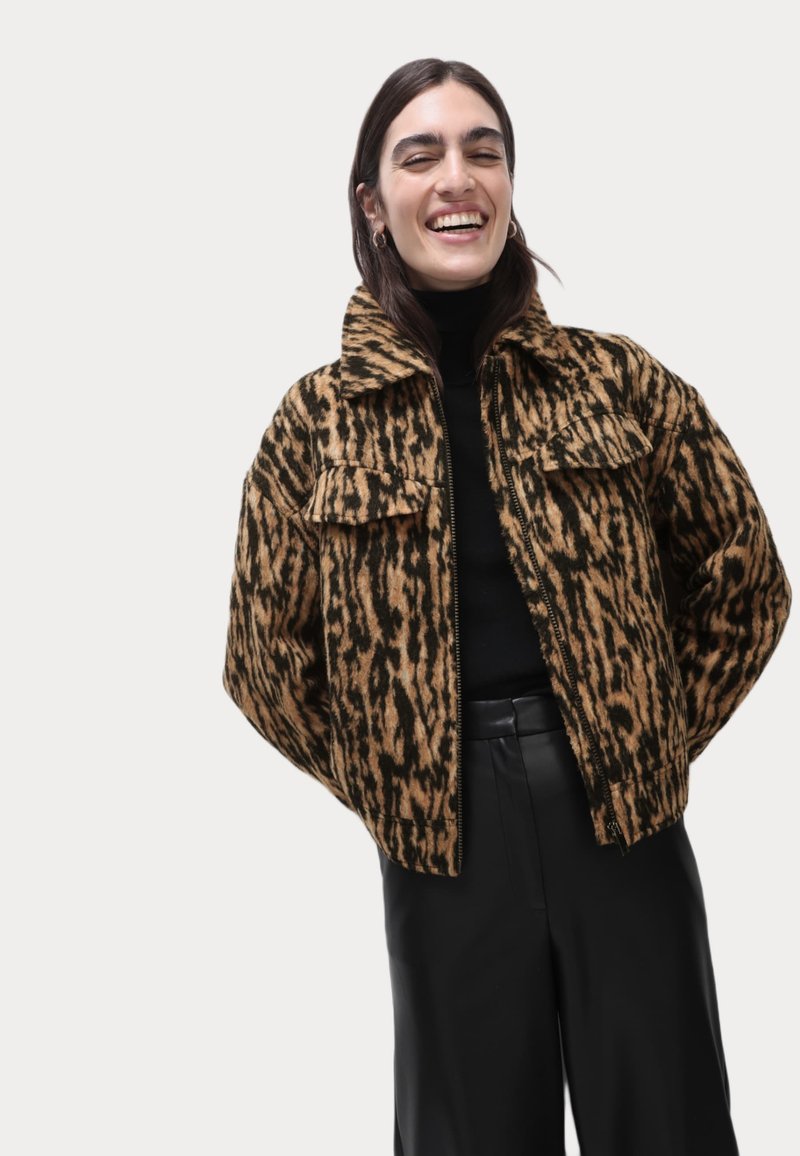 Veste à motifs marron et noir avec un col et des poches avant, portée sur un haut noir et associée à un pantalon noir.