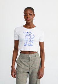 Hvid cropped t-shirt med blå grafisk print, der har tekst og en illustration. Bæres med ternede, højtaljede bukser. Glat tekstur.