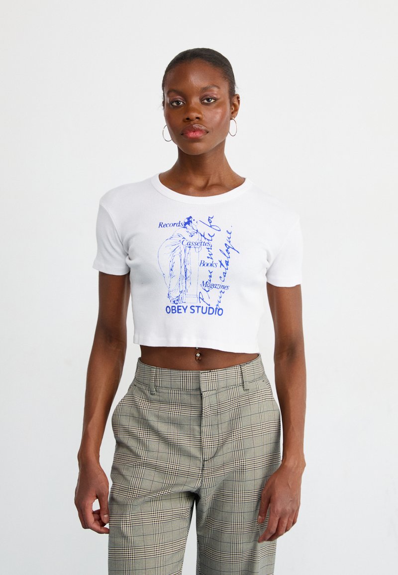Hvid cropped t-shirt med blå grafisk print, der har tekst og en illustration. Bæres med ternede, højtaljede bukser. Glat tekstur.