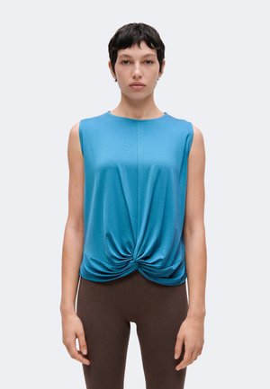 Jeune personne aux cheveux courts et foncés portant un haut bleu sans manches torsadé sur le devant et des leggings marron foncé, debout devant un fond uni.