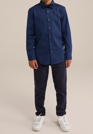 Bambino che indossa una camicia a maniche lunghe blu scuro con motivi, papillon nero, pantaloni scuri e scarpe da ginnastica bianche, in piedi davanti a uno sfondo semplice.