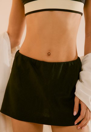 Nainen, jolla on musta crop-top ja lyhyt musta hame, valkoinen takki kevyesti hartioilla, paljastaen vatsanseutua.