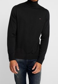 Suéter de cuello alto negro hecho de una tela suave, con mangas largas y un dobladillo ajustado. Pequeño logo bordado en el pecho.