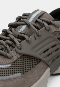Grau-brauner Sneaker aus Mesh- und Wildledermaterialien, mit Schnürsenkeln, reflektierenden Akzenten und einem dicken Sohledesign mit Ausschnitten an der Seite.