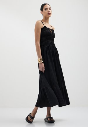 Schwarzes Maxikleid mit Rüsche-Trägern, gerüschtem Oberteil und gestuftem Rock. Kombiniert mit schwarzen Slippern und geschichteten metallic Armbändern.