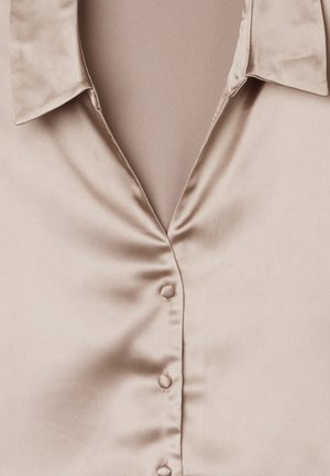 Blusa di seta di un leggero color beige, con colletto, chiusura frontale a bottoni e texture liscia.