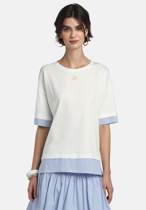 Vrouw draagt een wit shirt met korte mouwen over een blauw-wit gestreept shirt en een bijpassende geplooide rok, gecombineerd met creolen en armbanden als accessoires.