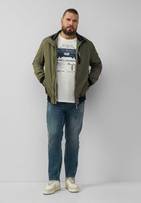 Olivgrüne Bomberjacke mit schwarzem Besatz, weißem Grafik-T-Shirt, blauen Jeans und beigen Turnschuhen, stehend vor einem schlichten Hintergrund.