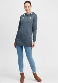 Felpa con cappuccio in tessuto blu scuro con accenti triangolari color sabbia, abbinata a jeans chiari strappati e stivaletti marroni alla caviglia.