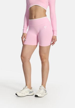 Donna che indossa un crop top a maniche lunghe rosa chiaro e pantaloncini da biker a vita alta con scarpe da ginnastica bianche su sfondo bianco.