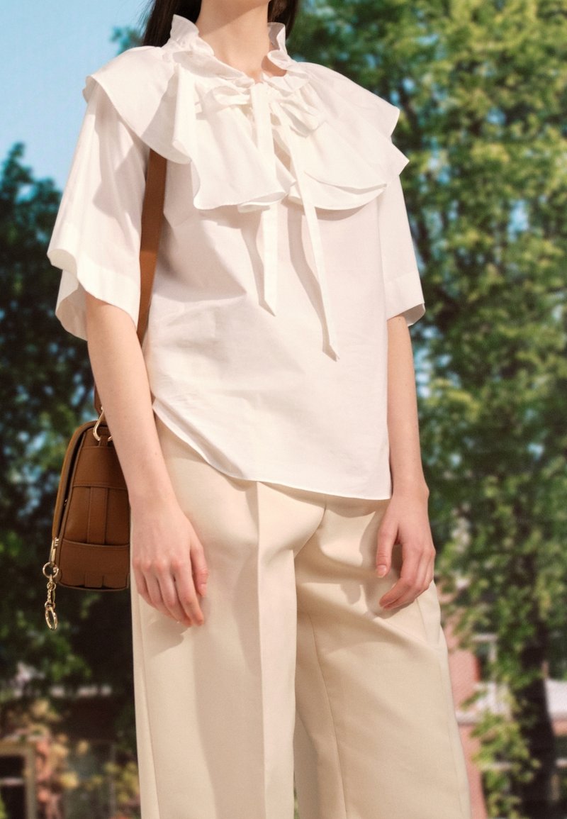 Blouse blanche avec grand col à volants et liens, associée à un pantalon large beige clair. Le mannequin tient un sac à bandoulière marron.