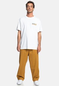 Hvid bomulds t-shirt med et logo i blå og gul, parret med løstsiddende sennepsgule bukser og hvide sneakers.