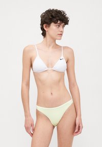 Balta trīsstūrveida bikini augšdaļa ar plāniem siksniņām, uz kuras redzams neliels melns logo. Apvienota ar gaiši dzeltenām bikini apakšdaļām, minimālistisks dizains.