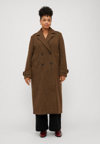 Cappotto lungo a quadri marrone con bottoni doppi, colletto a revers e tasche laterali. Abbinato a pantaloni neri a gamba larga e scarpe rosse.