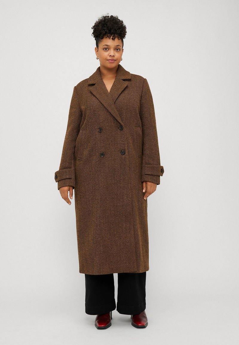Cappotto lungo a quadri marrone con bottoni doppi, colletto a revers e tasche laterali. Abbinato a pantaloni neri a gamba larga e scarpe rosse.
