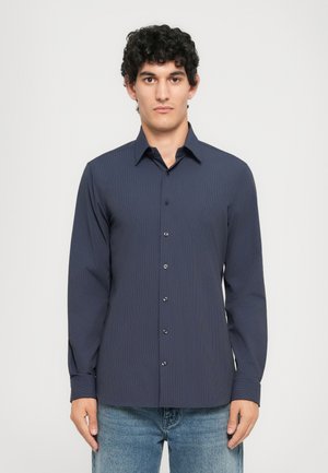 Mørkeblå skjorte med knappelukking og ribbet tekstur, lange ermer og svarte knapper. Brukt sammen med lyseblå jeans.