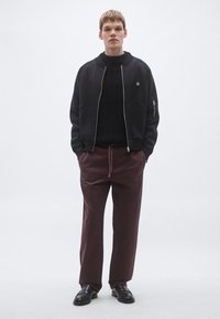 Geacă bomber neagră cu fermoar peste un pulover negru tricotat, asortată cu pantaloni bordo cu logo-ul „HUGO”. Pantofi negri din piele.