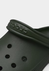 Close-up van een donker groene Crocs klomp met zichtbare textuur, ventilatiegaten en een verstelbare hielriem met ingegraveerde merknaam.
