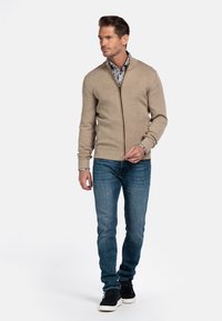 Beige cardigan met een ritssluiting, een gestreepte shirt eronder, gecombineerd met blauwe denim jeans en donkere sneakers. Gladde textuur en aansluitend ontwerp.