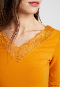 Long-sleeve top in orange cotton with a V-neck featuring floral lace trim in a slightly darker shade. Soft texture, fitted design.