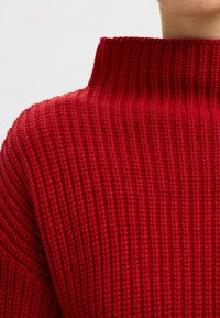 Roter Strickpullover mit hohem Kragen. Verfügt über eine gerippte Textur und ein grobes Strickmuster, das Wärme und Komfort betont.