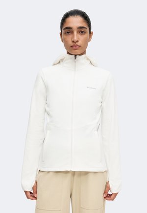 Femme aux cheveux foncés tirés en arrière, portant une veste blanche zippée avec capuche et un pantalon beige, debout devant un fond clair uni.
