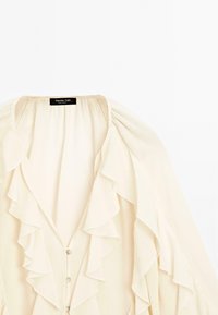 Blusa color crema con ruches a cascata sul fronte, caratterizzata da una profonda scollatura a V e chiusura con bottoni. Tessuto morbido e leggero.