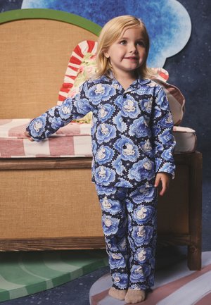 Noor laps sinistes mustrilistes pidzamaades seisab voodi kõrval, millel on triibulised padjad ja roheline kaarduv peats, öiste teemadega toas.