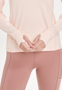 Hellrosa, langärmliges Sportoberteil mit Daumenlöchern, ausgestattet mit kontrastierenden Nähten. Kombiniert mit figurbetonten, pastellrosa Leggings.