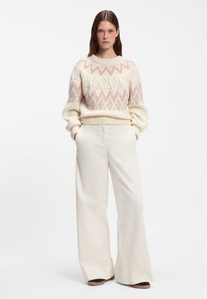 Kvinde med brunt hår iført en cremefarvet og lyserød mønstret sweater og hvide brede bukser, står med hænderne i lommerne på hvid baggrund.