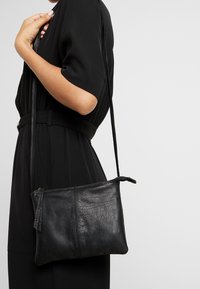 Borsa a tracolla in pelle nera con superficie structurata, chiusura con zip e tracolla sottile regolabile. Forma rettangolare semplice, senza motivi visibili.