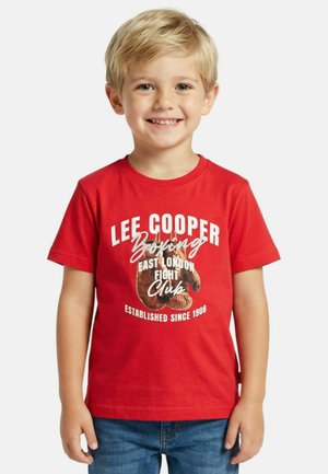Jeune garçon souriant aux cheveux blonds portant un t-shirt rouge Lee Cooper Boxing et un jean bleu, debout devant un fond clair uni.