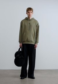 Χακί φούτερ με κουκούλα και μπροστινή τσέπη, συνδυασμένο με χαλαρά μαύρα παντελόνια. Το μοντέλο κρατάει μια στρογγυλή μαύρη τσάντα με χερούλι.