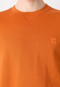 Gros plan sur un sweat-shirt orange à col rond avec un petit logo BOSS embossé sur le côté gauche de la poitrine et un détail de couture triangulaire sous le col.