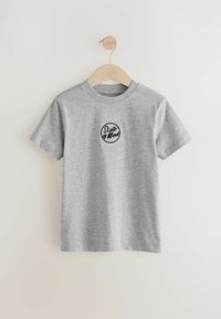 T-shirt di cotone grigio appesa a un gancio di legno, con un logo rotondo "Skate of Mind" in nero al centro. Maniche corte, girocollo.
