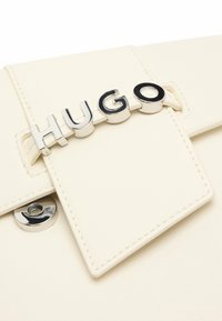 Valkoinen nahkainen käsilaukku, jossa on neliömäinen läppä ja krominen "HUGO" logo. Magneettinen napitussulku ja kontrastivärinen ompelu.