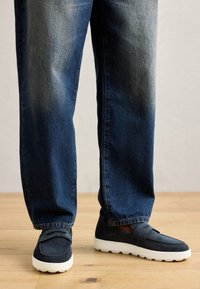 Marineblå semsket skinn slip-on sko med hvit gummisåle, kombinert med falmede blå rette jeans. Skoene har en enkel sydd detalj.