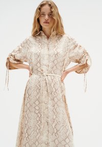 Beige napitna obleka s vzorcem kačjega usnja, s 3/4 rokavi, zavezovanjem na zapestjih in usklajenim pasom v pasu. Gladek tekstil.