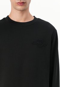 Schwarzer Pullover aus weichem Material mit Rundhalsausschnitt und gesticktem GANT-Logo in passender schwarzer Textur auf der Brust.