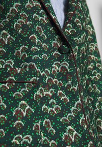 Blazer verde con pattern floreale bianco e bordeaux, dotato di colletto a punta e una tasca sul petto. Tessuto texturizzato con finitura lucida.